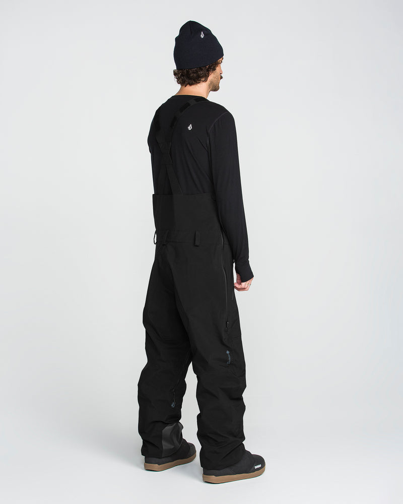 A person stands facing away, wearing Volcom Men’s Anyox 3L Gore Pro Bib snow pants with fully taped seams, along with a long-sleeve shirt, a beanie, and boots, against a plain light background.