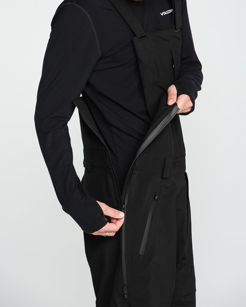 A person dressed in a long-sleeve black shirt unzips the side of the Volcom Men’s Anyox 3L Gore Pro Bib, black overalls made from recycled-nylon Pro Shell with a chest pocket. The background is plain white.