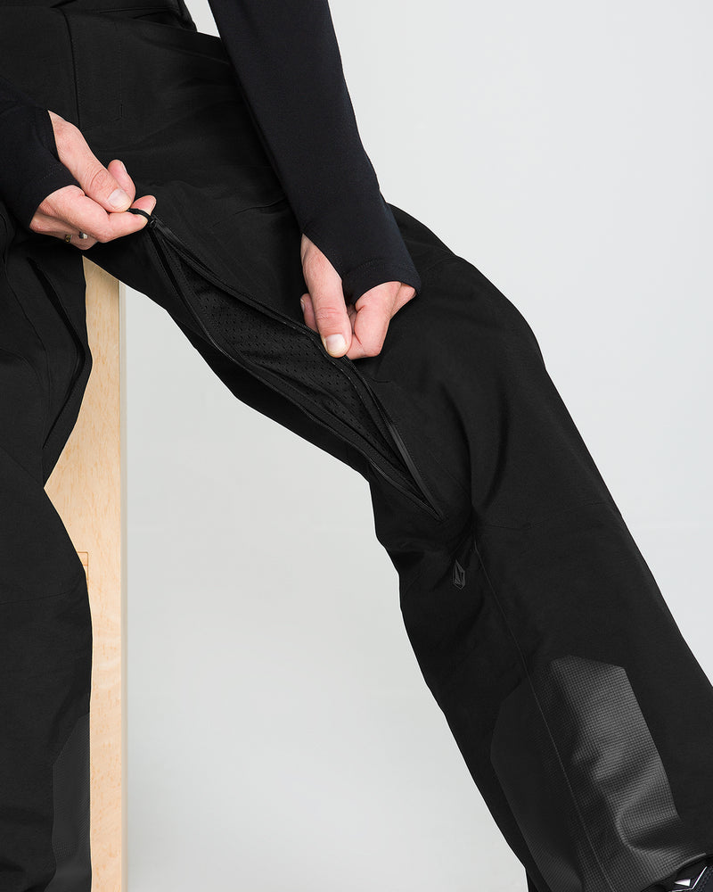 A person sitting on a wooden stool unzips a vent on the side of their black Men’s Anyox 3L Gore Pro Bib from Volcom, revealing a mesh lining inside—crafted from recycled-nylon Pro Shell with fully taped seams for added durability.