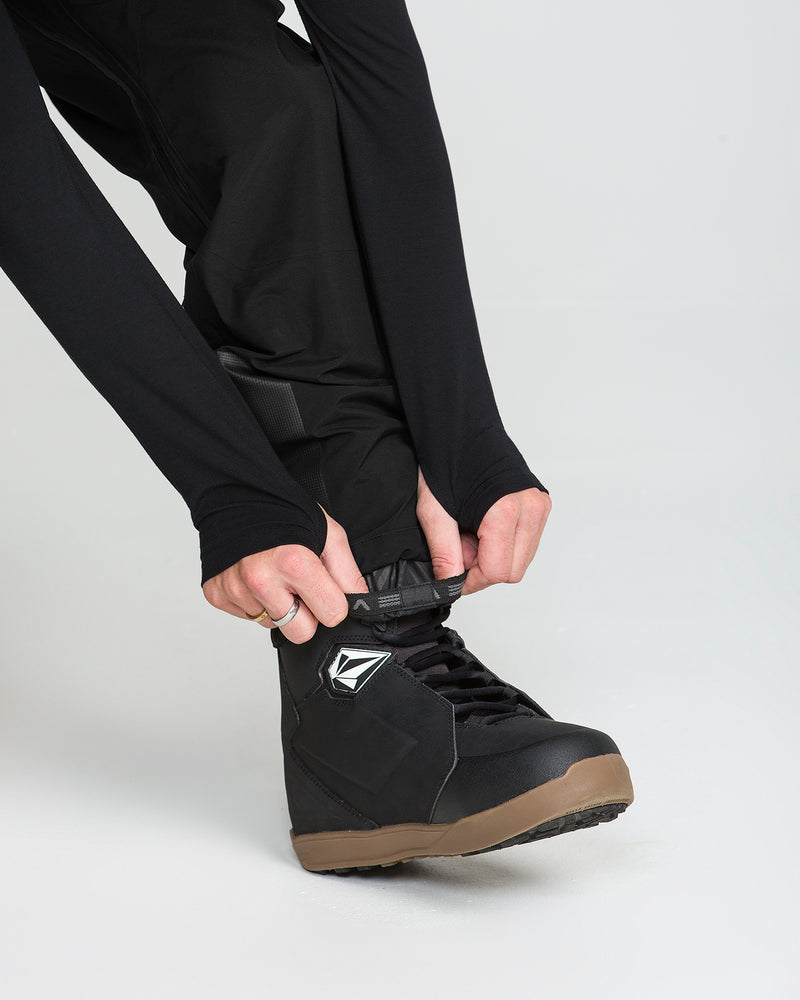 A person wearing the Volcom Men’s Anyox 3L Gore Pro Bib—constructed with recycled-nylon Pro Shell material and fully taped seams—pulls on a black snowboarding boot with a tan sole against a plain light background.