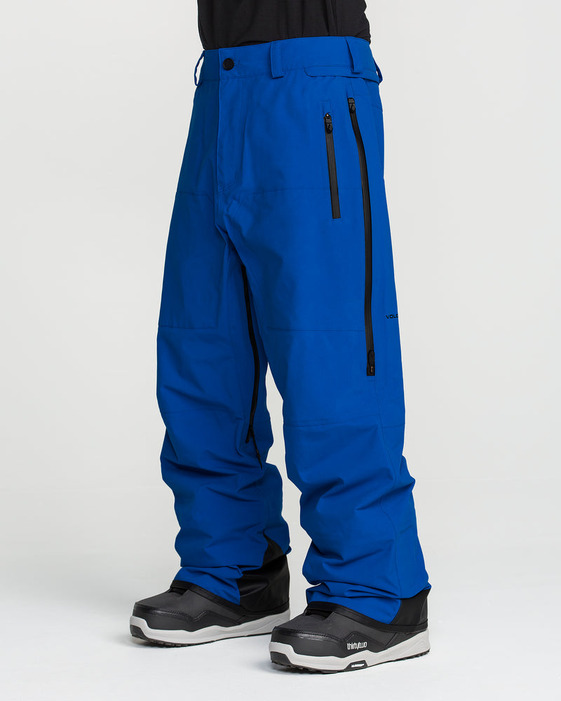 A person wearing Volcom Men’s Guch 3L Gore-Tex Pro Pants in bright blue with black zippers and black and gray snow boots, standing against a plain white background.