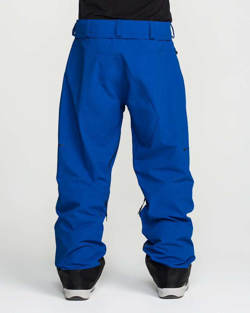 A person is shown from the back wearing Volcom Men’s Guch 3L Gore-Tex Pro Pants in bright blue and black boots. The pants feature a relaxed fit, large back pocket, belt loops, and are PFAS-free. The background is plain white.