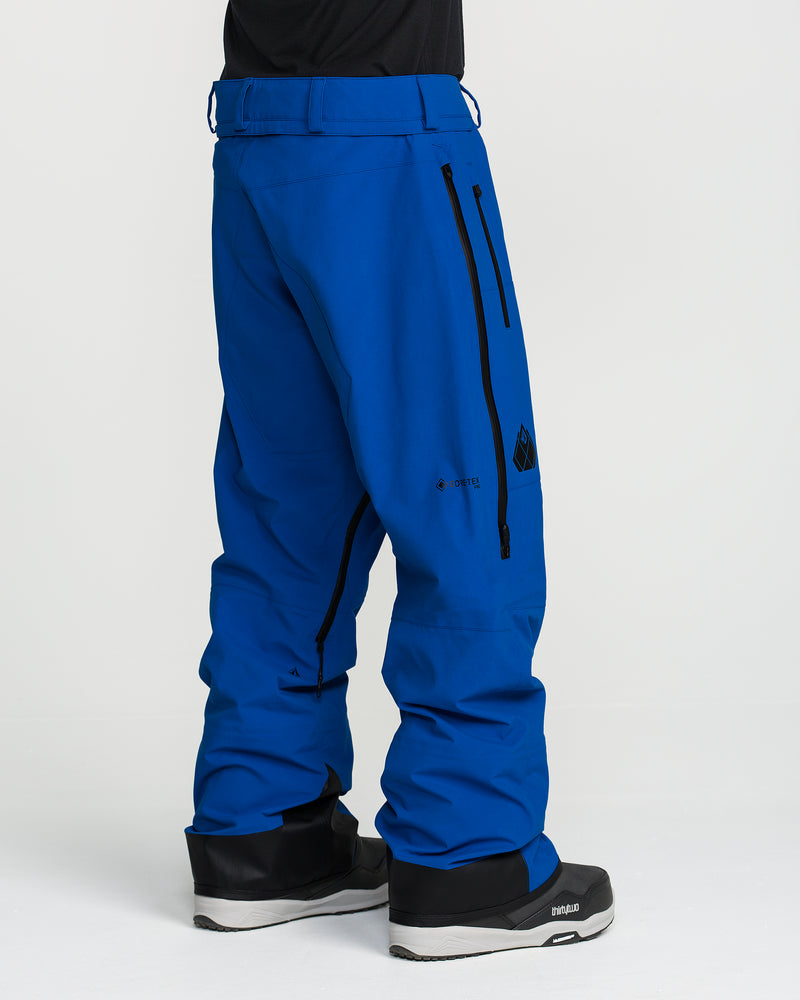 A person wearing Volcom Men’s Guch 3L Gore-Tex Pro Pants in bright blue with black zipper details and black snow boots, standing against a plain white background.