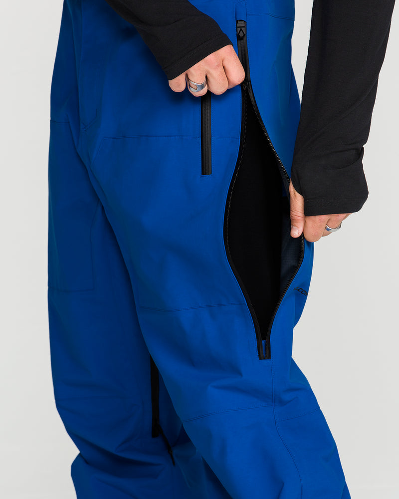 A person wearing bright blue Volcom Men’s Guch 3L Gore-Tex Pro Pants and a black long-sleeve shirt unzips a large side zipper on the pants, revealing a black inner lining.