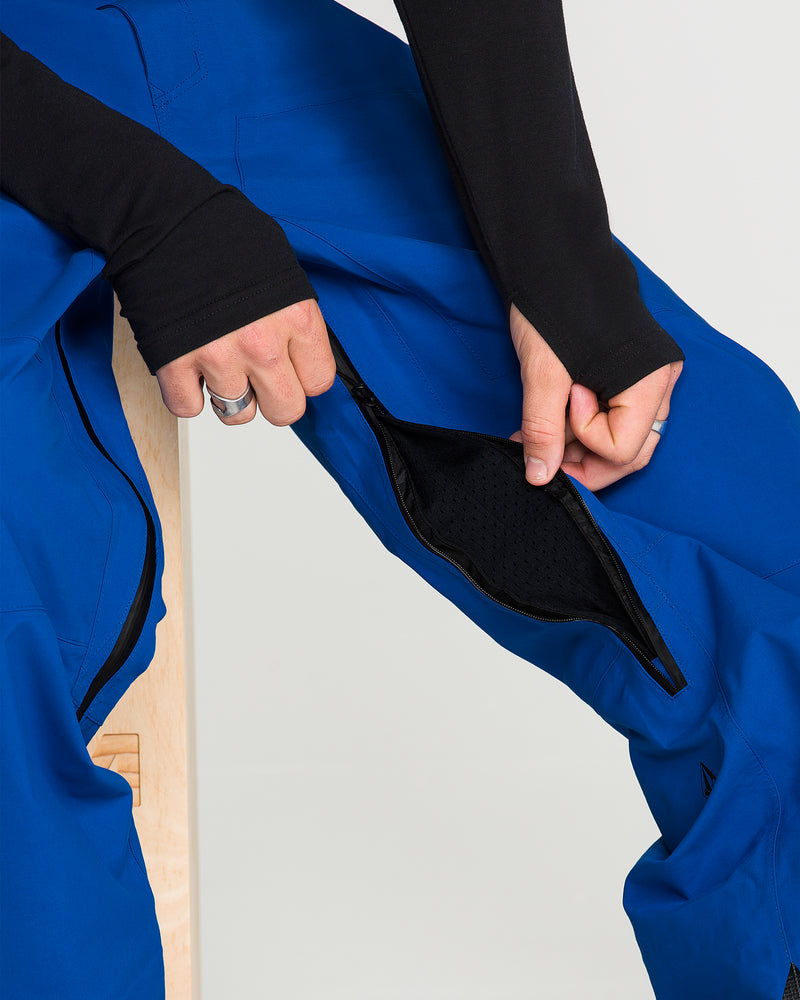 Seated on a stool, a person unzips the black mesh-lined ventilation opening of bright blue Volcom Men’s Guch 3L GORE-TEX Pro Pants, revealing the inner fabric. The person is wearing a long-sleeve black top and silver rings.