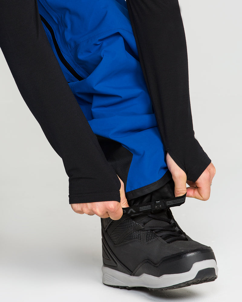 A person wearing Volcom Men’s Guch 3L Gore-Tex Pro Pants in black and blue with black shoes is adjusting an ankle strap. Only their hands, lower legs, and shoes are visible.