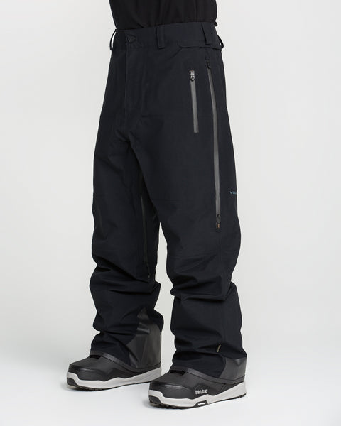 A person wearing black Volcom Men’s Guch 3L Gore-Tex Pro Pants, which are PFAS-free for advanced protection and sustainability, featuring waterproof construction and zipper pockets, paired with black snow boots. The image is cropped above the waist and has a plain white background.