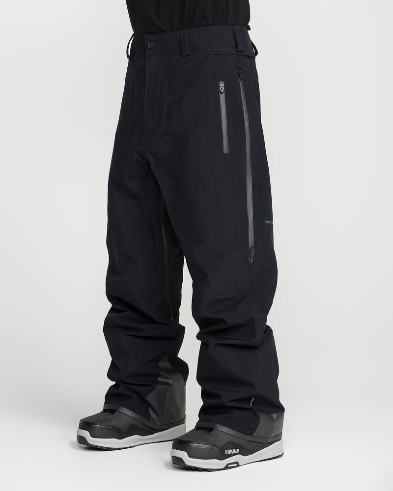 A person wearing black Volcom Men’s Guch 3L Gore-Tex Pro Pants, which are PFAS-free for advanced protection and sustainability, featuring waterproof construction and zipper pockets, paired with black snow boots. The image is cropped above the waist and has a plain white background.