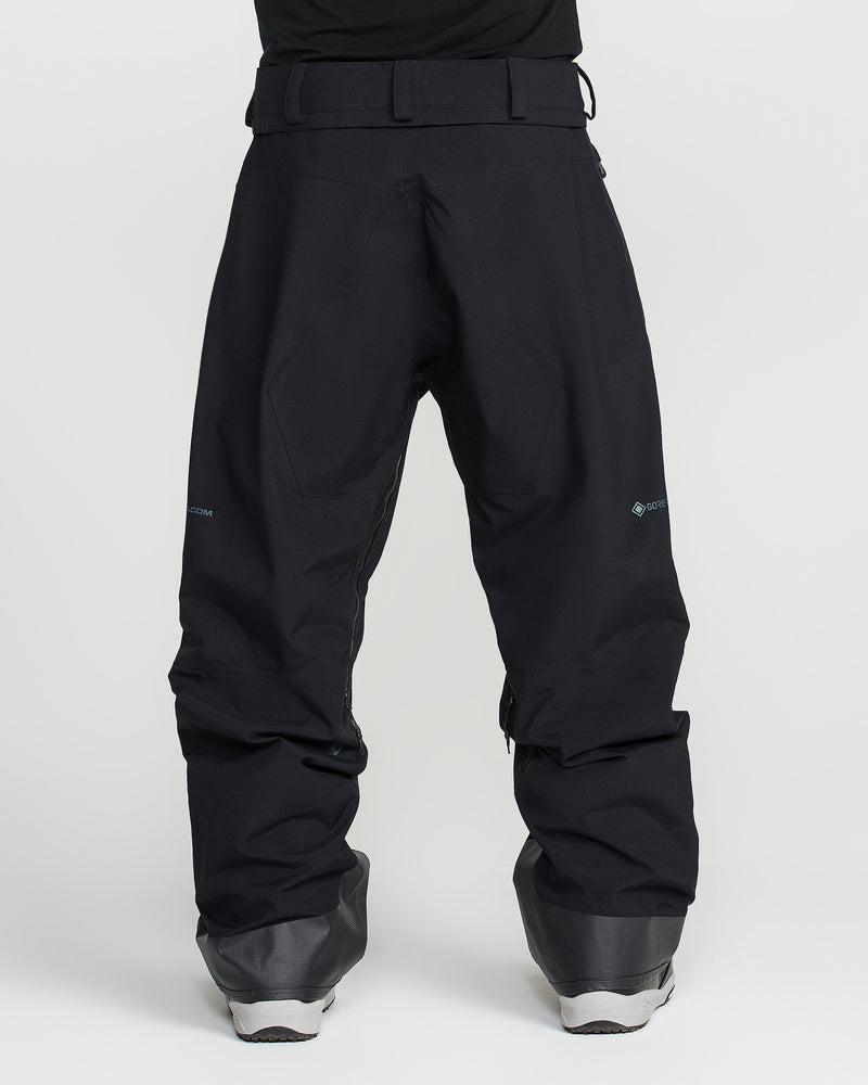 A person wearing loose-fitting black Volcom Men’s Guch 3L Gore-Tex Pro Pants and gray snow boots stands facing away from the camera against a plain white background.