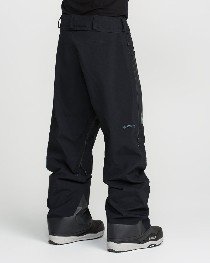 A person wearing black Volcom Men’s Guch 3L GORE-TEX Pro Pants and black snow boots stands on a white background, facing away from the camera. The pants feature zippered pockets, RECCO technology, and display Gore-Tex branding.