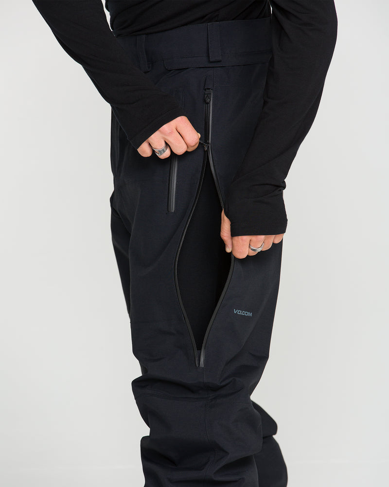 A person wearing black Men’s Guch 3L GORE-TEX Pro Pants uses their left hand to unzip a thigh pocket. The PFAS-free Volcom pants display the brand name on the upper leg, with only the lower torso and legs visible.