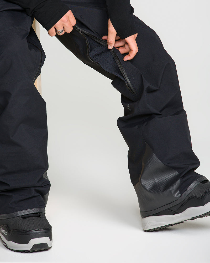 A person opens a zippered vent on the inner thigh of Volcom Men’s Guch 3L Gore-Tex Pro Pants, revealing the mesh lining, while wearing black winter boots.