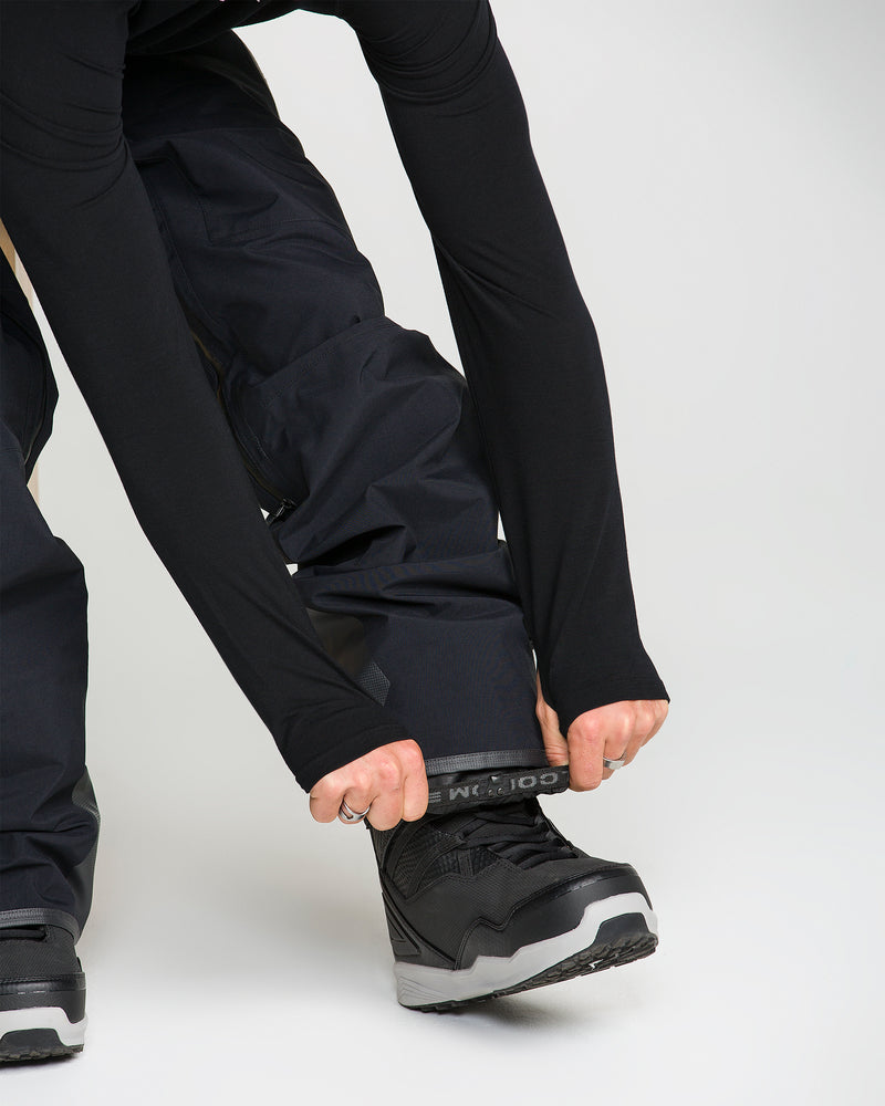 A person wearing black Volcom Men’s Guch 3L Gore-Tex Pro Pants and black shoes pulls an elastic gaiter over their ankle, preparing for outdoor or winter activities in these PFAS-free, eco-friendly pants.