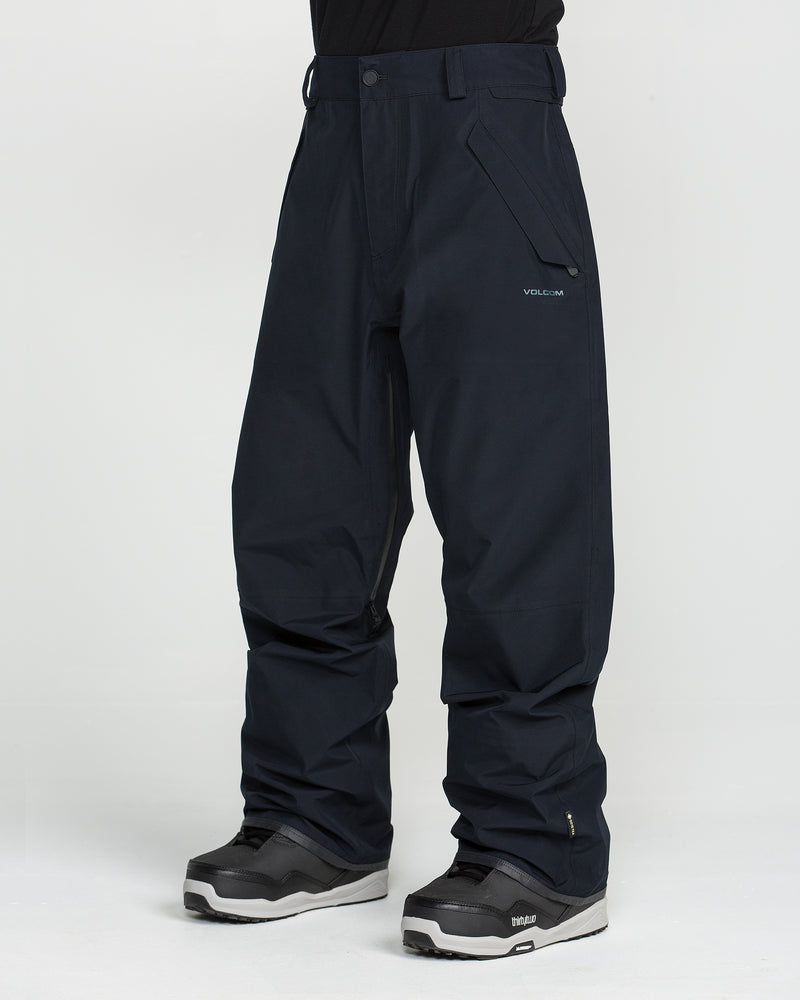 A person is shown from the waist down, standing against a plain white background, wearing black Volcom Unisex Tester 3L Gore-Tex Pants with visible side pockets and a Volcom logo, paired with black snow boots.