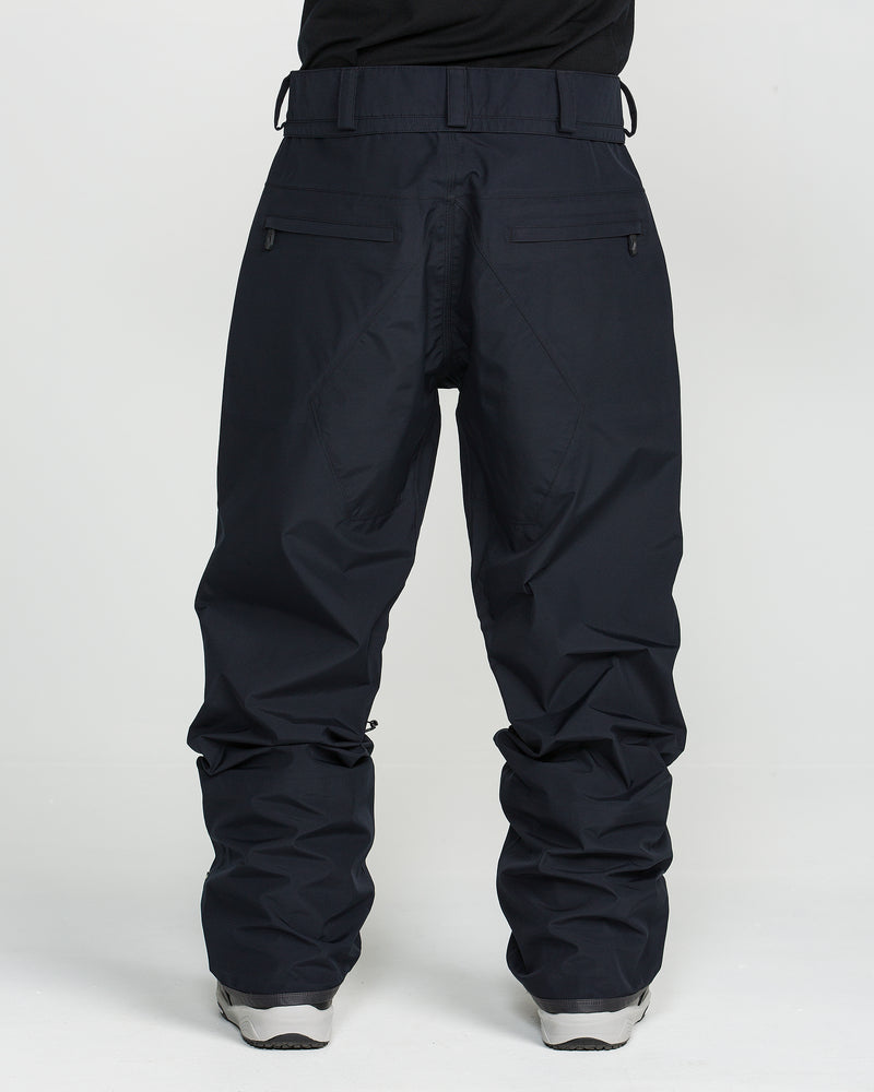 A person is seen from the back wearing Volcom Unisex Tester 3L Gore-Tex Pants in a loose-fitting, dark navy design, featuring zippered back pockets and belt loops for added functionality. The outfit is paired with light gray sneakers and set against a plain white background.