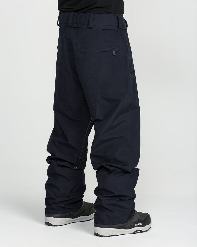 A person wearing loose-fitting, dark-colored Volcom Unisex Tester 3L Gore-Tex Pants and black winter boots stands against a plain white background, facing away from the camera.