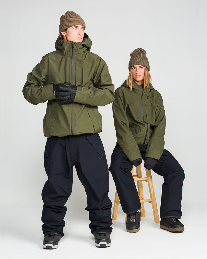 Two people in olive green jackets, brown knit beanies, and gloves pose in a studio against a white background. Both wear winter boots and the Volcom Unisex Tester 3L Gore-Tex Pants in black—one stands with arms crossed while the other sits on a wooden stool.