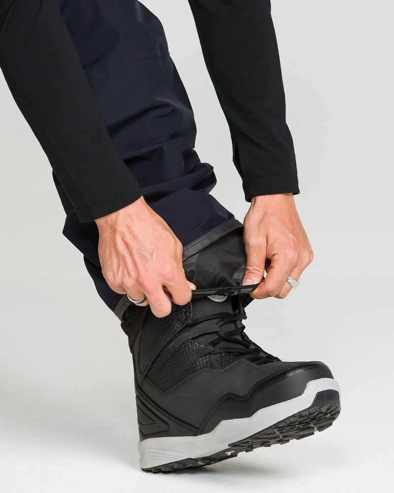 A person wearing Volcom Unisex Tester 3L Gore-Tex Pants and boots adjusts a gaiter over their boot, securing it in place with their hands. Their waterproof snow pants from Volcom offer reliable protection against wet conditions during outdoor adventures.