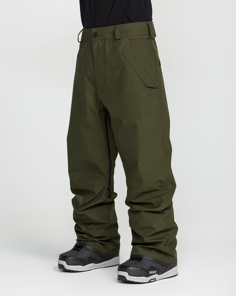 A person wearing loose-fitting olive green Volcom Unisex Tester 3L Gore-Tex Pants and black snow boots stands against a plain white background, with only the lower half of their body visible.