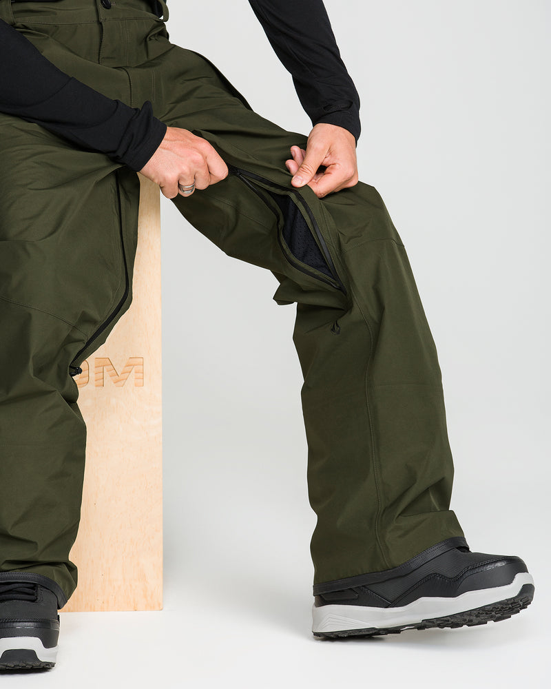 A person unzips the side vent of their olive green Volcom Unisex Tester 3L Gore-Tex Pants while sitting on a light wooden stool, wearing black long sleeves and gray-and-black shoes.