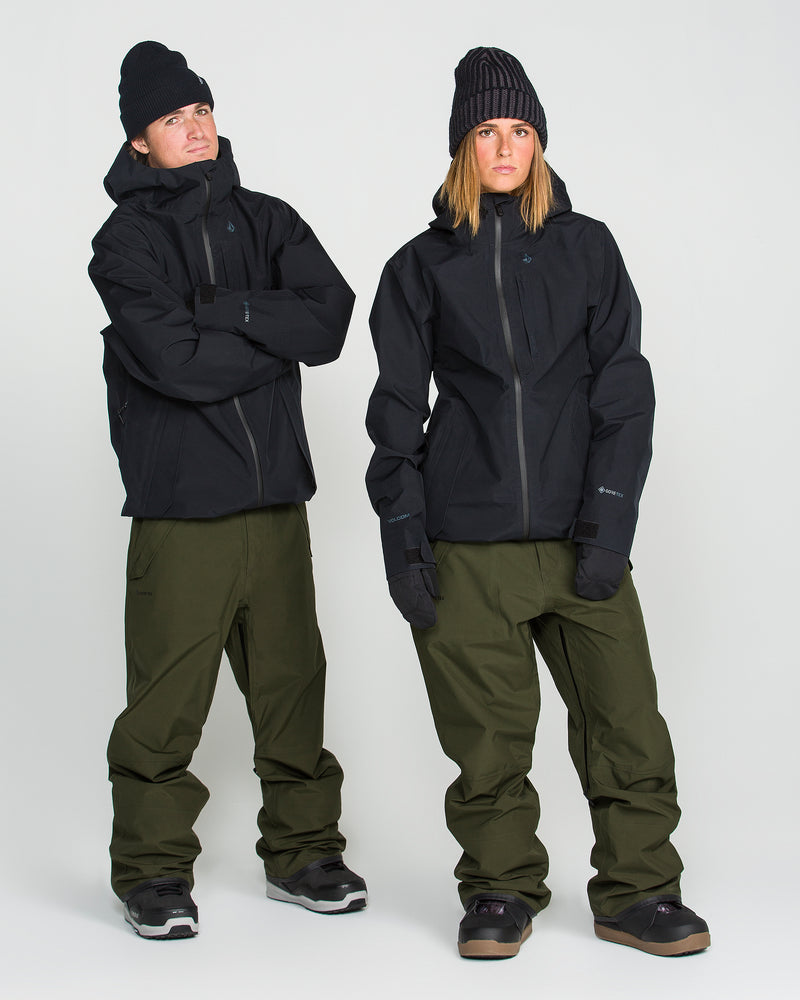 Two people in black jackets, Volcom Unisex Tester 3L Gore-Tex Pants in olive green, and black beanies stand side by side against a plain white background, both looking at the camera with neutral expressions.