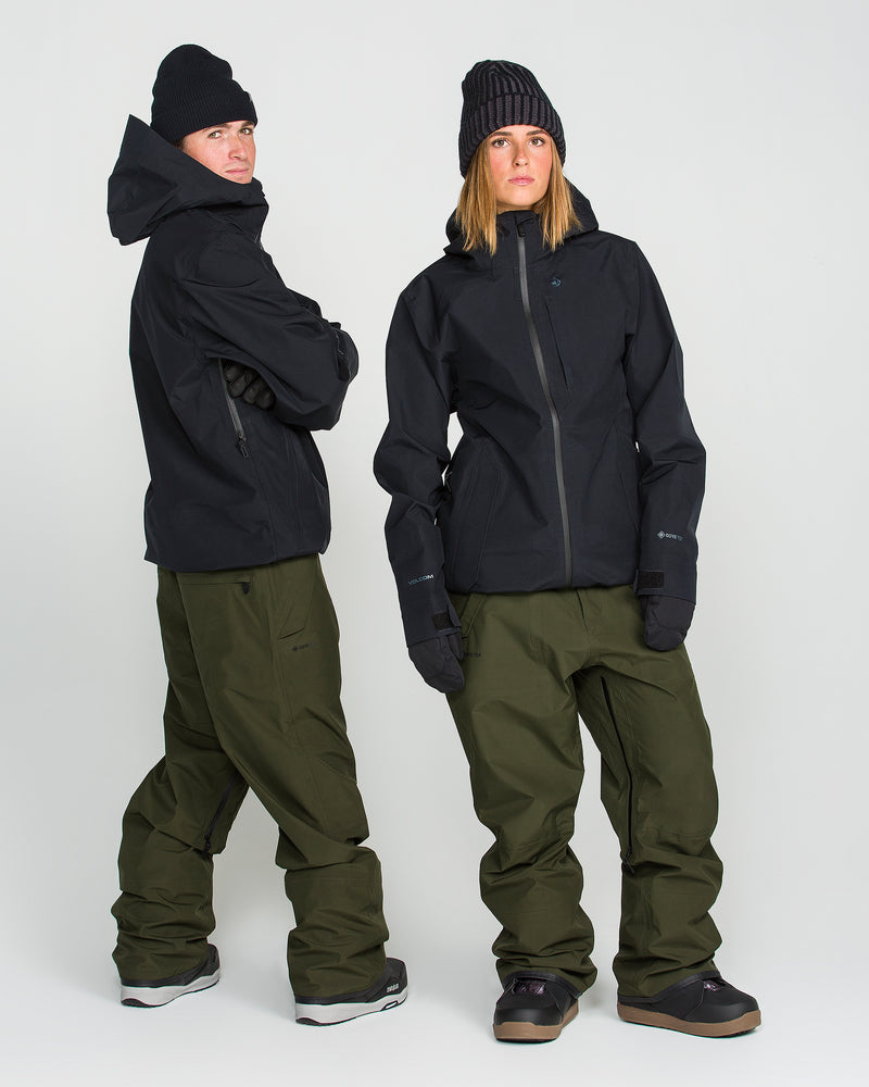 Two people stand against a white background, facing different directions and modeling winter outerwear; both are wearing black jackets, black knit hats, black gloves, and olive green Volcom Unisex Tester 3L Gore-Tex Pants made from recycled materials.