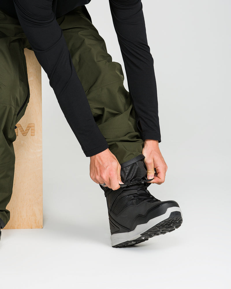 A person wearing green Volcom Unisex Tester 3L Gore-Tex Pants and a black long-sleeve shirt adjusts their black and gray boot while sitting on a wooden bench against a plain background.