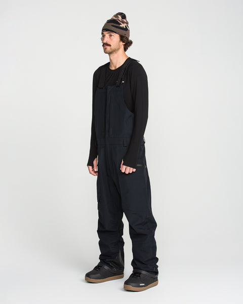 A person wearing a black long-sleeve shirt, Volcom Men’s Rain GORE-TEX Bib overall, black shoes, and a camouflage beanie stands against a plain white background.