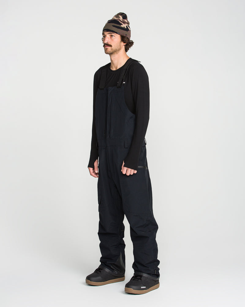 A person wearing a black long-sleeve shirt, Volcom Men’s Rain GORE-TEX Bib overall, black shoes, and a camouflage beanie stands against a plain white background.