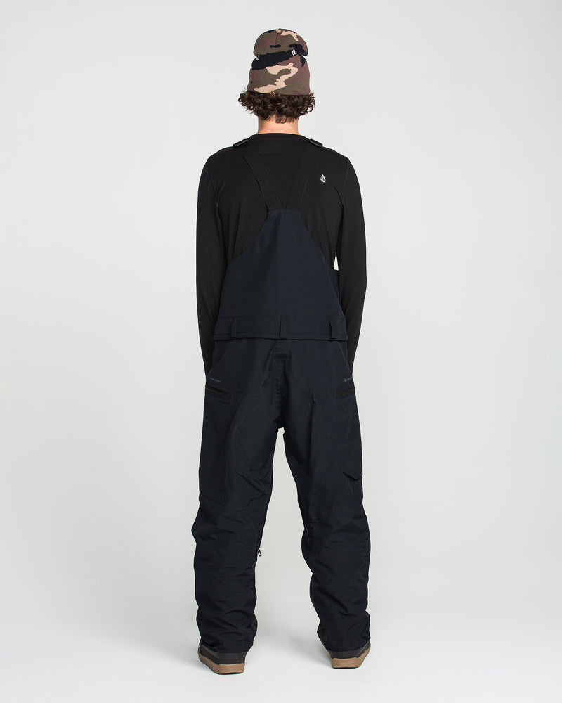 A person stands facing away, wearing a black long-sleeve shirt, loose Volcom Men’s Rain GORE-TEX Bib overalls, and a pink and brown camouflage beanie, against a plain white background.