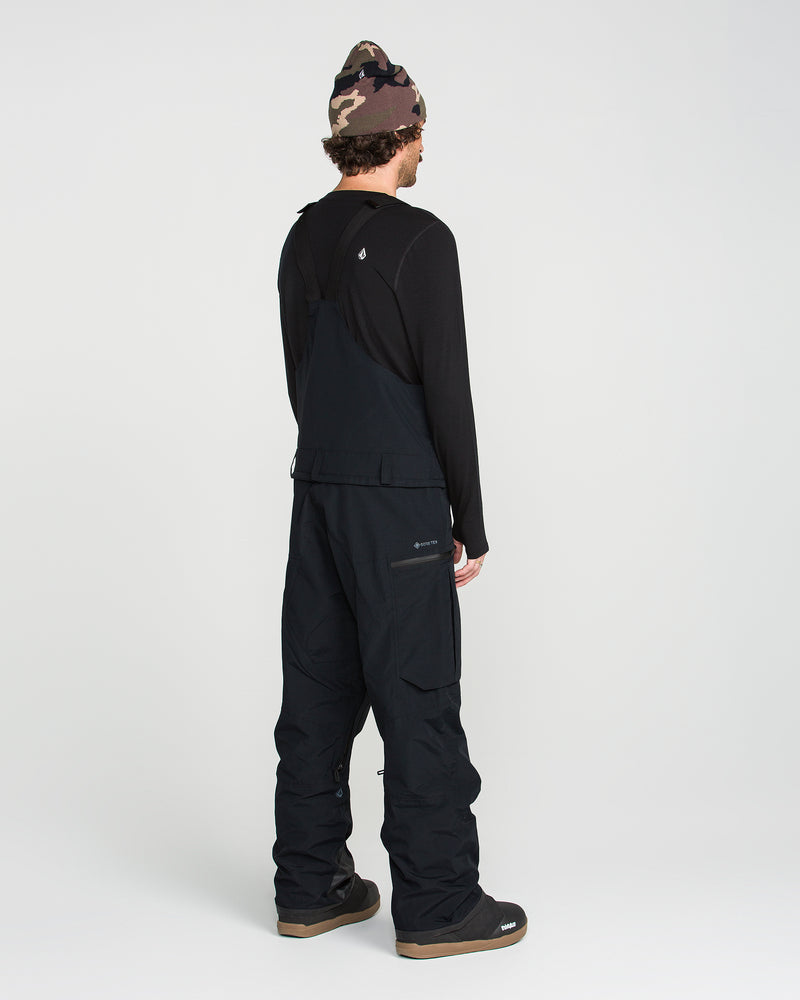 A person stands facing away, wearing Volcom Men’s Rain Gore-Tex Bib overalls in black, paired with a black long-sleeve shirt, black boots, and a camouflage beanie, on a plain light background.
