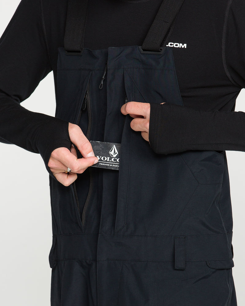 A person, shown from the torso down against a plain background, wears the Volcom Men’s Rain Gore-Tex Bib over a long-sleeve shirt and inserts a Volcom-branded tag into the zippered chest pocket.
