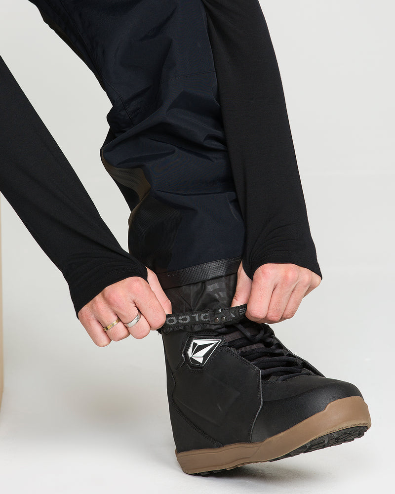 A person wearing black Volcom Men’s Rain Gore-Tex Bib snow pants and a long-sleeve shirt fastens the strap on a black and brown snowboard boot.