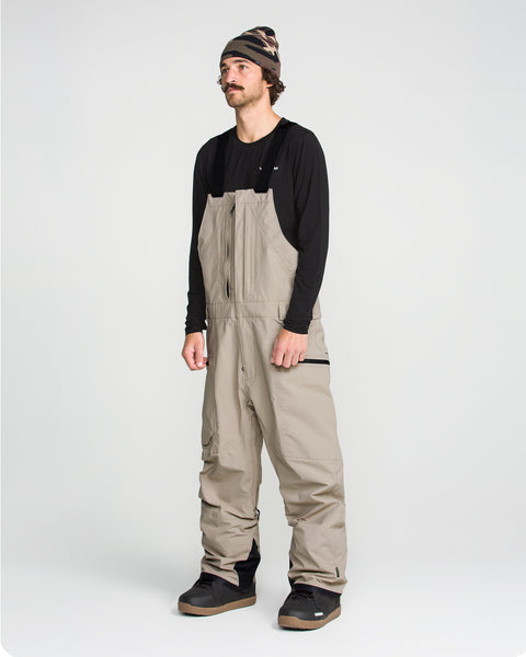 A person wearing a black long-sleeve shirt, Volcom Men’s Rain Gore-Tex Bib overalls made from recycled waterproof materials with a breathable lining, dark boots, and a striped beanie stands against a plain light background.