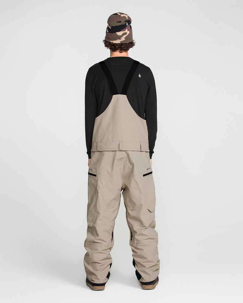 A person stands facing away, wearing the Volcom Men’s Rain Gore-Tex Bib in tan with breathable lining, a black long-sleeve shirt, a brown camouflage beanie, and brown boots, set against a plain white background.