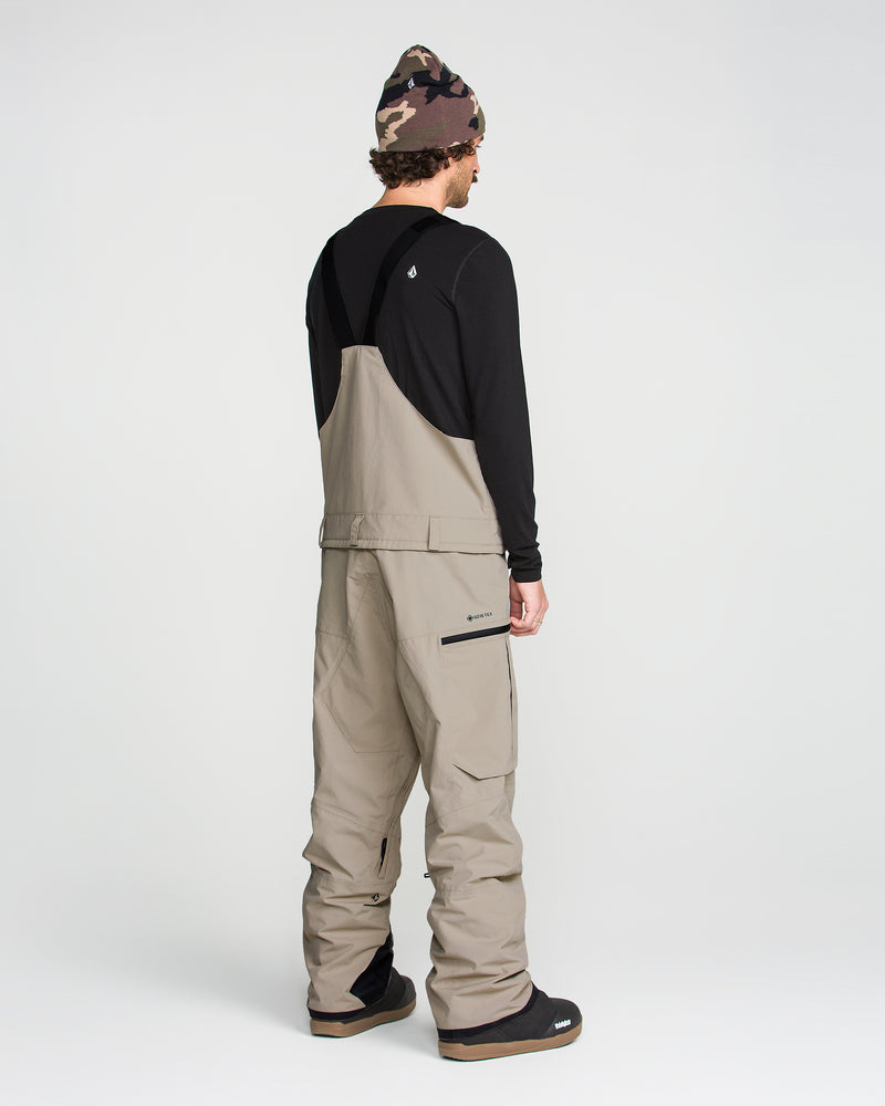 A person is seen from behind against a plain light background, wearing the Volcom Men’s Rain Gore-Tex Bib in beige, which features a breathable lining, paired with a black long-sleeve shirt, black boots, and a brown camouflage beanie.