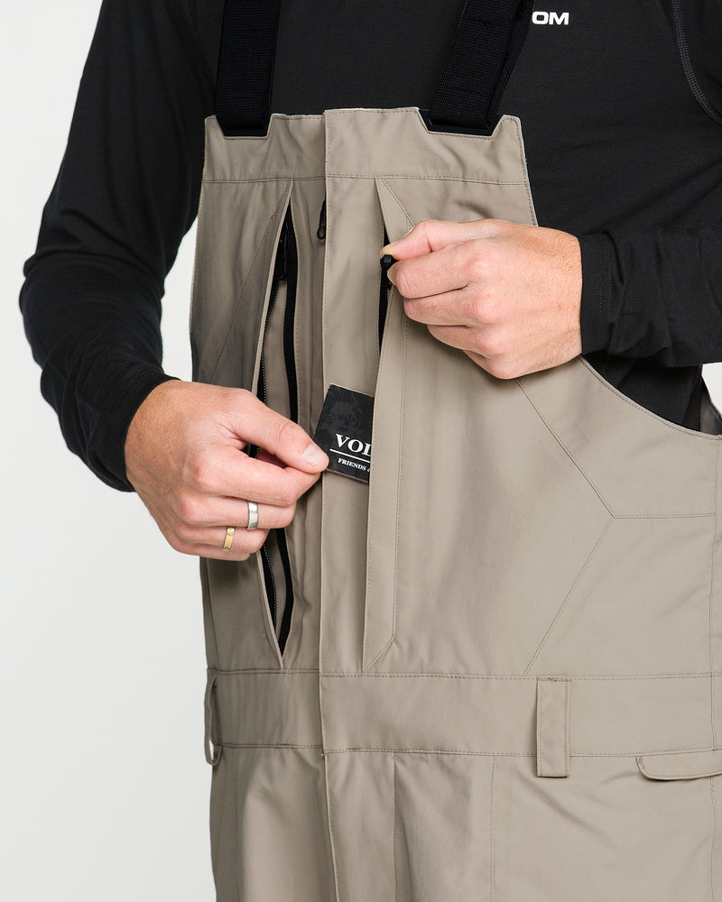 A person wearing the Volcom Men’s Rain Gore-Tex Bib over a black shirt unzips a chest pocket to reveal a wallet inside.