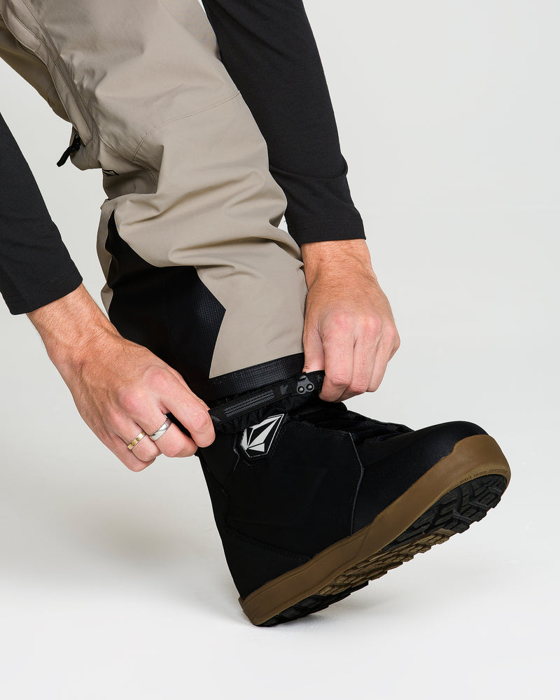 A person wearing the Volcom Men’s Rain Gore-Tex Bib pairs tan pants with black boots, fastening the boot gaiter over the top of the boot with both hands. Only their lower legs and hands are visible against a white background, showcasing the bib’s breathable lining and GORE-TEX recycled materials engineered by Volcom for comfort and weather protection.
