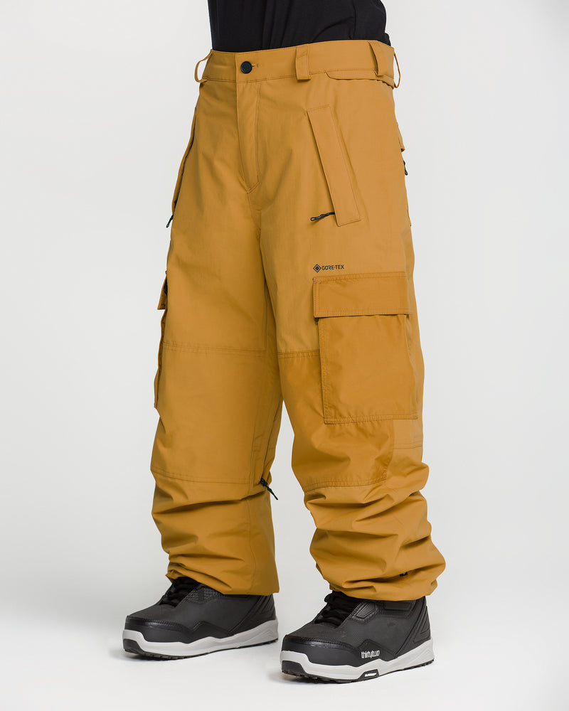 A person is wearing loose-fitting mustard yellow Volcom Men’s Longo Gore-Tex Pants with multiple pockets and black snow boots, standing against a plain white background. Inspired by Arthur Longo, these pants offer both style and functionality for the slopes.