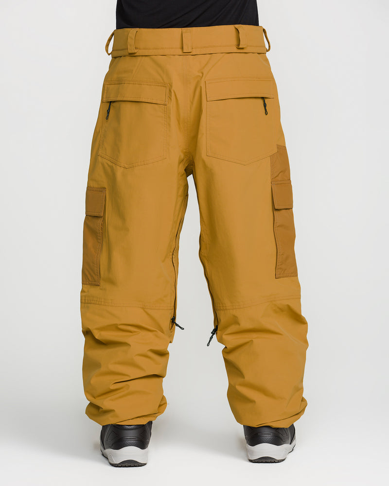A person wears Volcom Men’s Longo GORE-TEX Pants in mustard yellow—a baggy-fit snowboard style inspired by Arthur Longo—paired with black shoes and photographed from the back against a plain white background. These pants feature large pockets, zippers, and reinforced sections on the sides.