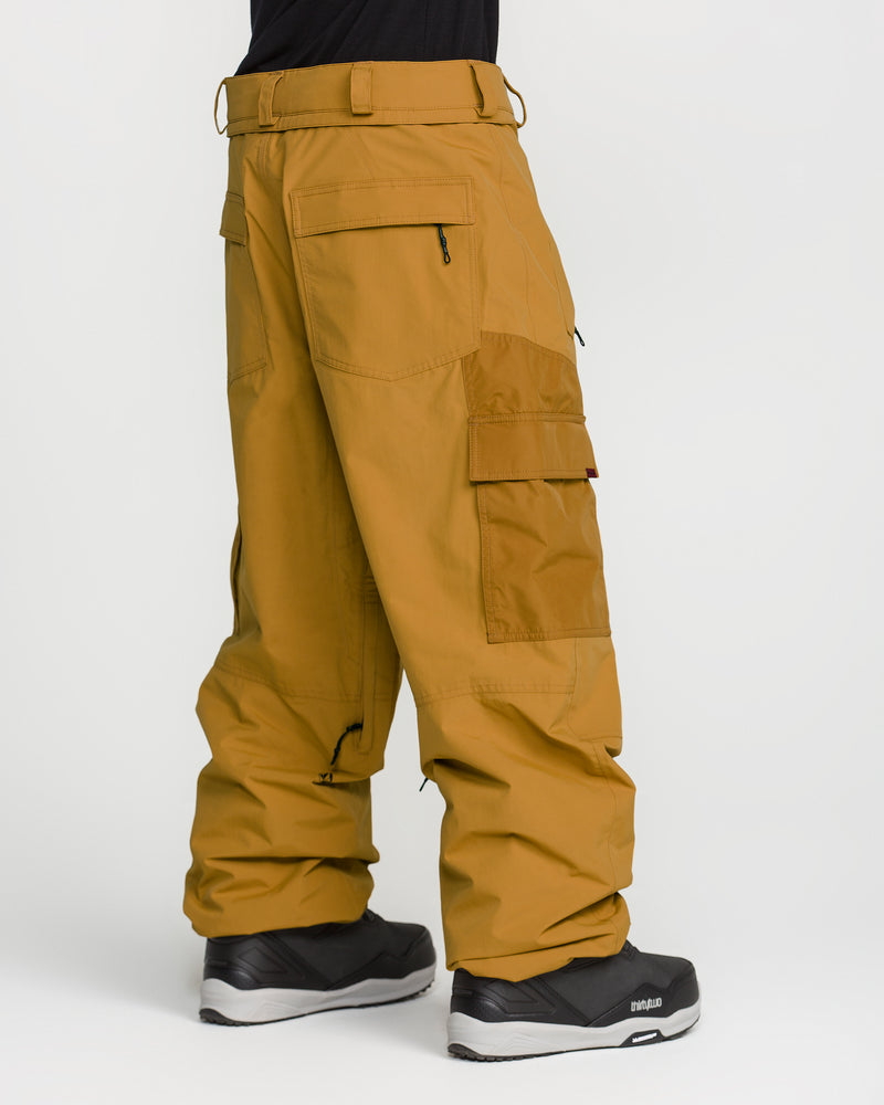 A person is shown from the side and back against a plain white background wearing the Volcom Men’s Longo Gore-Tex Pants in mustard yellow with a baggy fit, inspired by Arthur Longo, featuring multiple zippered pockets, paired with black athletic shoes.