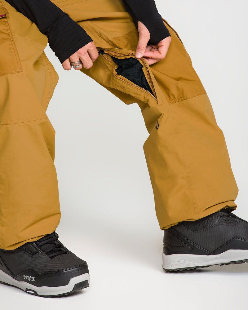 A person wearing Volcom Men’s Longo GORE-TEX Pants in mustard yellow and black boots crouches to unzip the inner thigh vent of the pants. Their hands are visible against a plain white background.