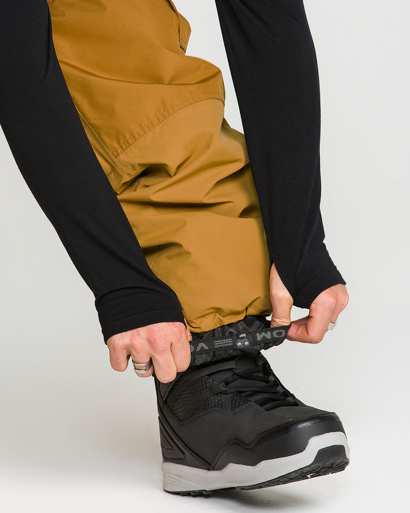 A person wearing black boots and mustard yellow Volcom Men’s Longo Gore-Tex Pants adjusts the pants over the top of their boot. Only their lower legs and hands are visible, echoing Arthur Longo’s signature style.