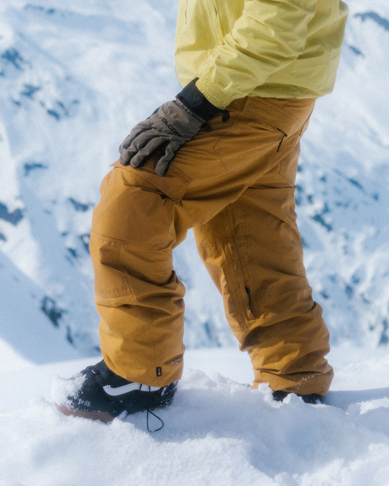 Dressed in Volcom Men’s Longo Gore-Tex Pants, a person stands on snow amid a backdrop of snowy mountains, with only the lower half of their body—clad in premium snowboarding gear—visible.