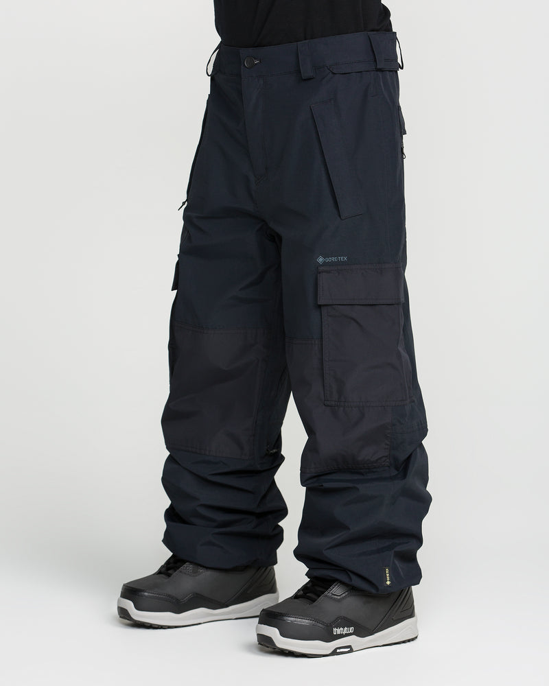 A person is wearing black Volcom Men’s Longo Gore-Tex Pants—cargo snow pants inspired by Arthur Longo’s signature baggy fit, designed with multiple pockets and reinforced knee areas—paired with black and gray snowboard boots, standing against a plain white background.
