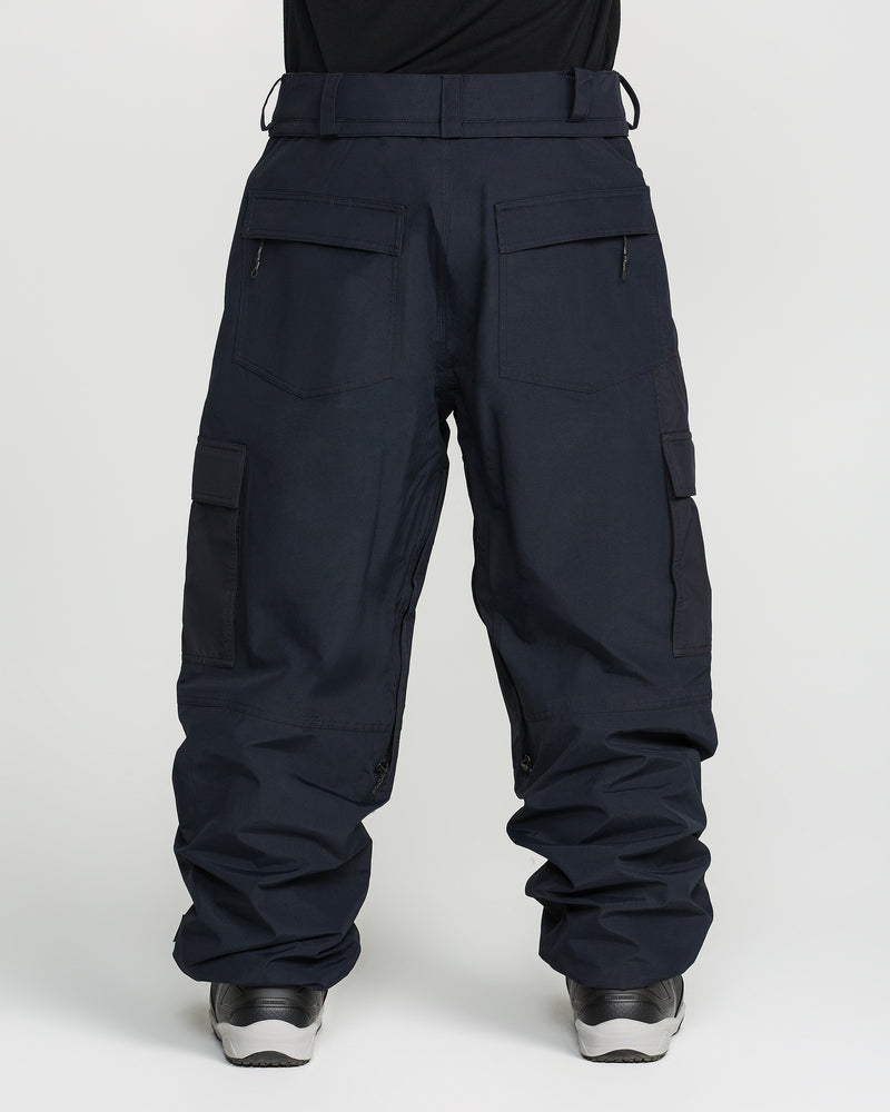 A person is shown from the back waist down, dressed in Volcom’s Men’s Longo Gore-Tex Pants—loose-fitting dark cargo pants inspired by Arthur Longo’s signature baggy-fit snowboard style, featuring multiple pockets—paired with a black shirt tucked in and gray athletic shoes, standing against a plain light background.