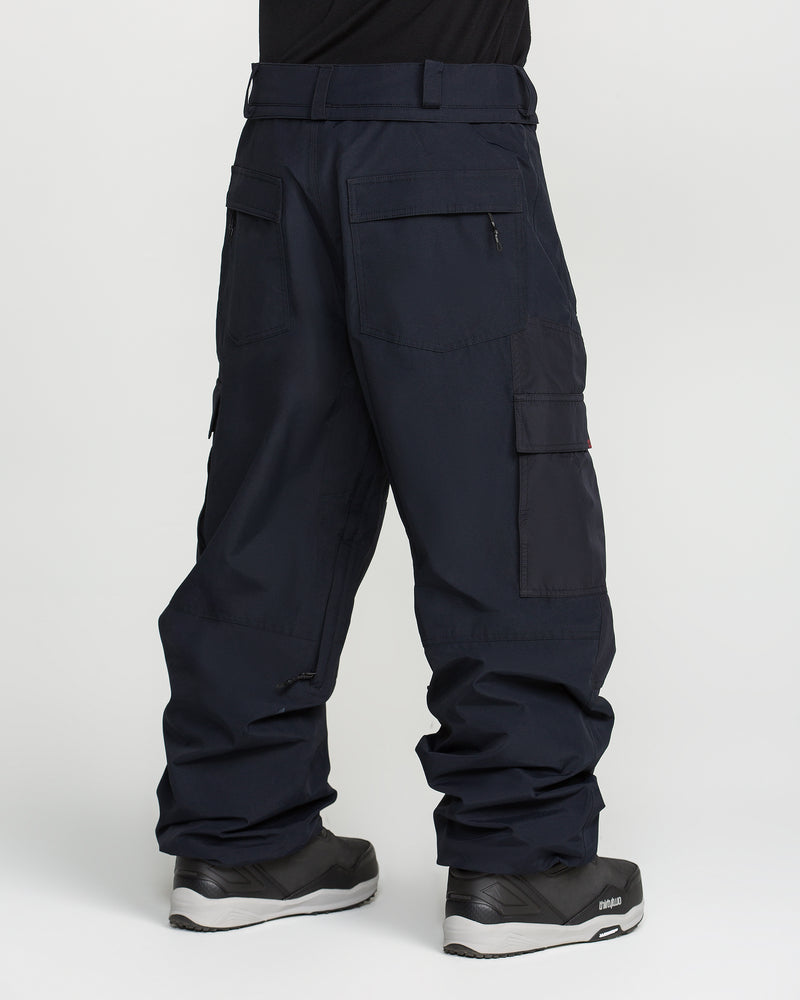 A person wearing Volcom Men’s Longo Gore-Tex Pants—a pair of loose-fitting dark cargo pants reminiscent of baggy-fit snowboard pants—paired with black boots, stands with their back to the camera against a plain white background.
