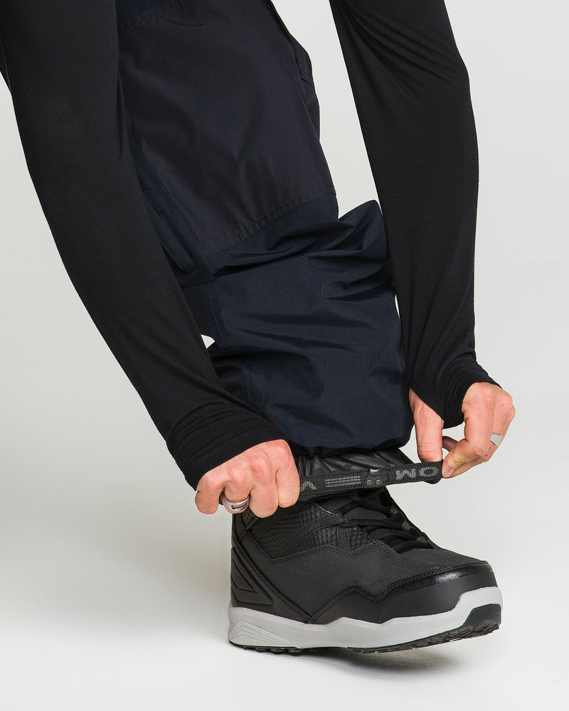 Wearing Volcom Men’s Longo Gore-Tex Pants, a person adjusts the strap at the cuff of their baggy-fit snowboard pants over their black shoes, channeling Arthur Longo’s signature style. The GORE-TEX pants stand out against a plain, light background.