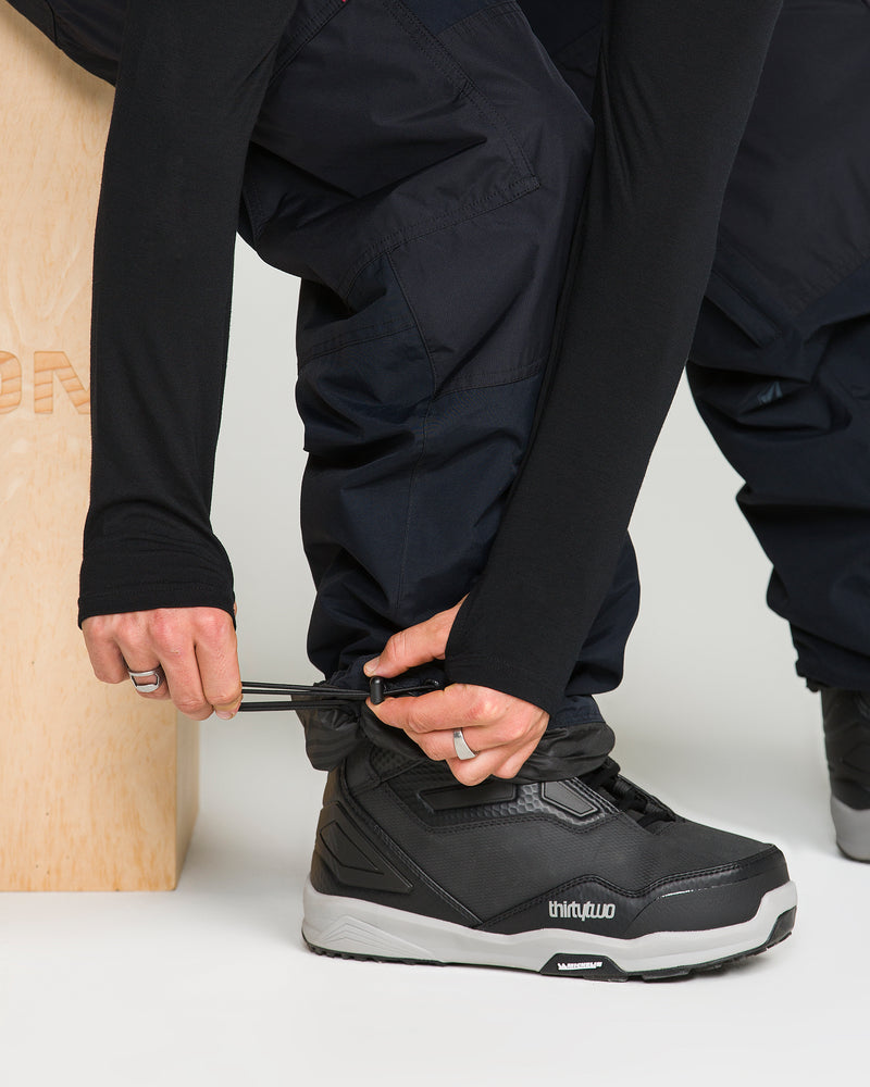 A person wearing Volcom Men’s Longo Gore-Tex Pants in a black, baggy fit squats to tighten the ankle cuff over a black snowboard boot using the built-in drawstring. A wooden box is partially visible on the left.