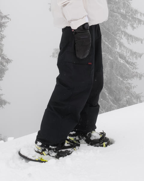 A person wearing black Volcom Men’s Longo Gore-Tex Pants, a white jacket, and black gloves stands in deep snow with snowshoes. Snow-covered trees and foggy weather can be seen in the background.